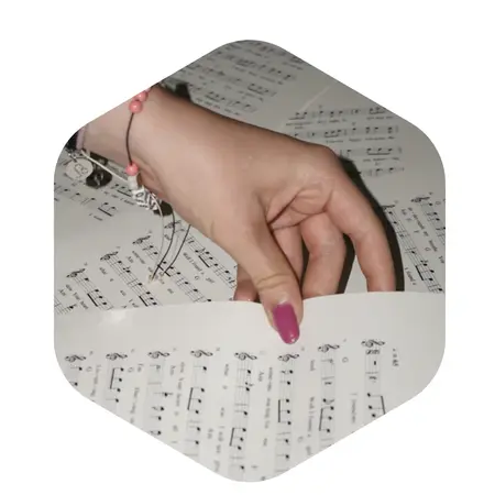 Eine Hand blättert durch Notenblätter, die auf einem Tisch liegen. Die Noten sind deutlich sichtbar, und die Hand hat lange, mit Nagellack lackierte Fingernägel. Die Umgebung ist unscharf, sodass der Fokus auf dem Blättern der Noten liegt.