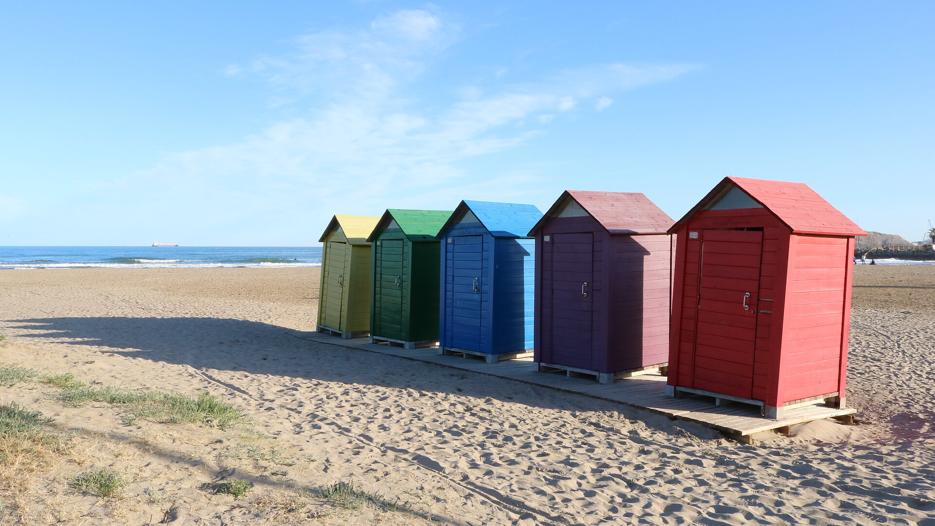 Strand mit Meer im Hintergrund ist sichtbar. In der Mitte des Bildes sind 5 verschiedenfarbige kleine Strandhäuschen sichtbar