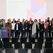 Eine große Gruppe von Menschen steht zusammen auf der Bühne, um an der Veranstaltung "2. Wiesbadener Tag für sozial nachhaltiges Investieren in Immobilien" teilzunehmen. Im Hintergrund ist ein Präsentationsbildschirm zu sehen.