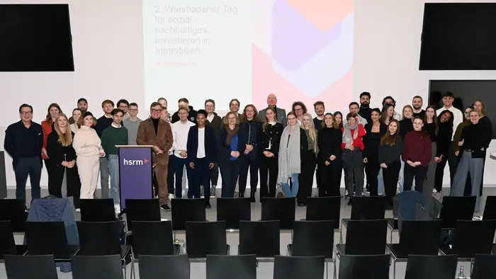 Eine große Gruppe von Menschen steht zusammen auf der Bühne, um an der Veranstaltung "2. Wiesbadener Tag für sozial nachhaltiges Investieren in Immobilien" teilzunehmen. Im Hintergrund ist ein Präsentationsbildschirm zu sehen.