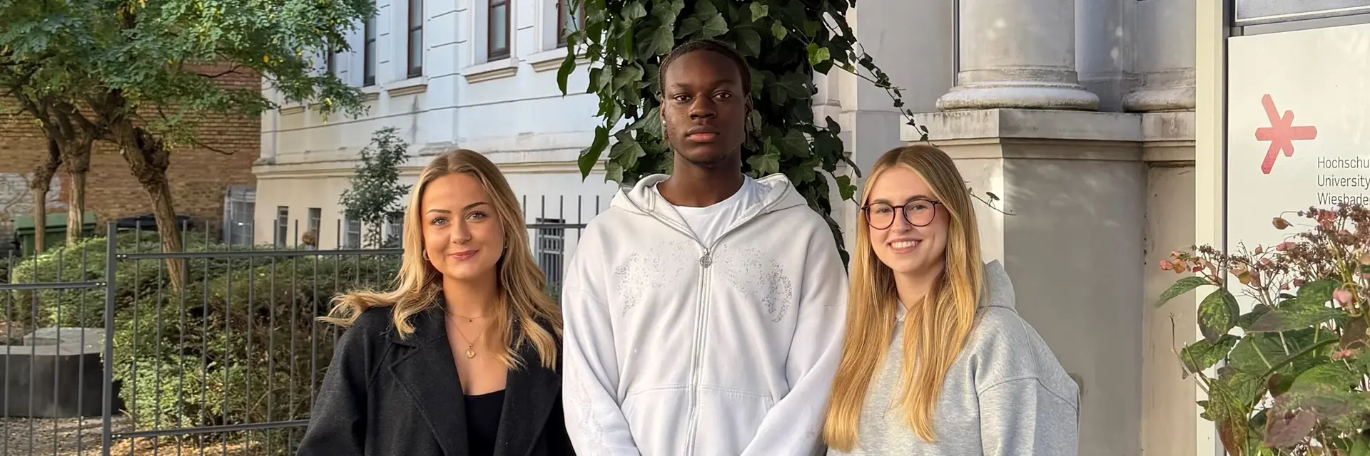 Drei junge Menschen stehen nebeneinander im Freien, umgeben von Bäumen und Gebäuden. Sie lächeln freundlich in die Kamera. Die Person links trägt einen schwarzen Mantel, die mittlere Person ein weißes Sweatshirt und die rechts eine graue Sweater mit blauer Jeans.