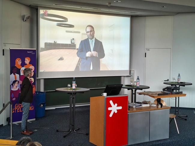 Ein Mann in einem Anzug steht auf einem Bildschirm und spricht. Er hat eine Brille und ist in einem modernen Raum mit einem Tisch und zwei Stühlen sichtbar. Im Vordergrund ist eine Person zu sehen, die ihm gegenübersteht.