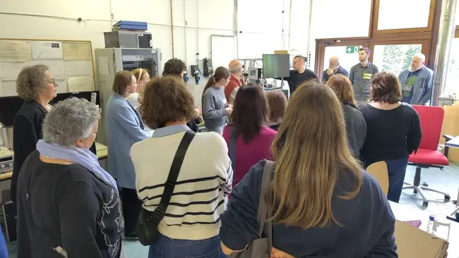 Eine Gruppe von Menschen steht in einem Raum und hört einem Vortrag zu. Einige Teilnehmer sind in Gespräche vertieft, während andere aufmerksam nach vorne schauen. Der Raum ist hell erleuchtet und enthält technische Geräte und Möbel.