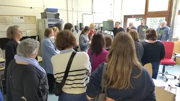 Eine Gruppe von Menschen steht in einem Raum und hört einem Vortrag zu. Einige Teilnehmer sind in Gespräche vertieft, während andere aufmerksam nach vorne schauen. Der Raum ist hell erleuchtet und enthält technische Geräte und Möbel.