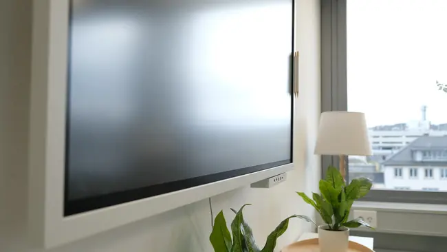 Das Bild zeigt einen modernen Flachbildfernseher, der an der Wand montiert ist. Rechts neben dem Fernseher befindet sich ein Fenster mit Blick nach draußen. Darunter steht ein kleiner Tisch mit einer Lampe und einer Pflanze in einem Topf.