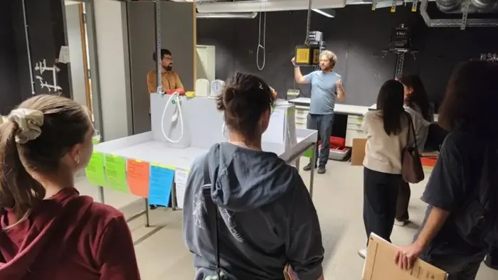 Eine Gruppe von Personen steht in einem Raum und hört einem Redner zu, der an einem Tisch steht. Auf dem Tisch befinden sich verschiedene Geräte. Bunte Zettel hängen an der Wand, und die Atmosphäre wirkt informativ und aufmerksam.