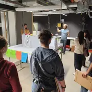 Eine Gruppe von Personen steht in einem Raum und hört einem Redner zu, der an einem Tisch steht. Auf dem Tisch befinden sich verschiedene Geräte. Bunte Zettel hängen an der Wand, und die Atmosphäre wirkt informativ und aufmerksam.