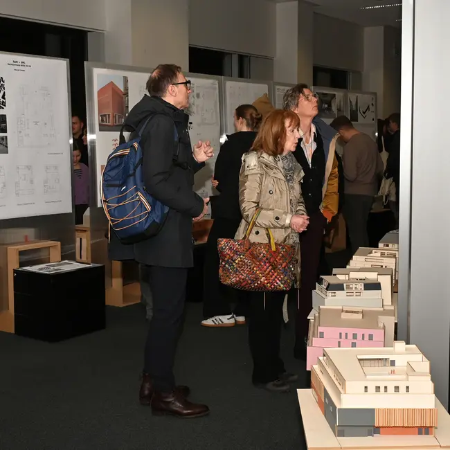 Man sieht eine Gruppe von Menschen, die an der Ausstellung der Abschlussarbeiten im Studiengang Architektur teilnehmen. Sie betrachten mehrere Modellgebäude, die auf Tischen ausgestellt sind. Im Hintergrund sind Plakate mit architektonischen Zeichnungen oder Plänen zu sehen.
