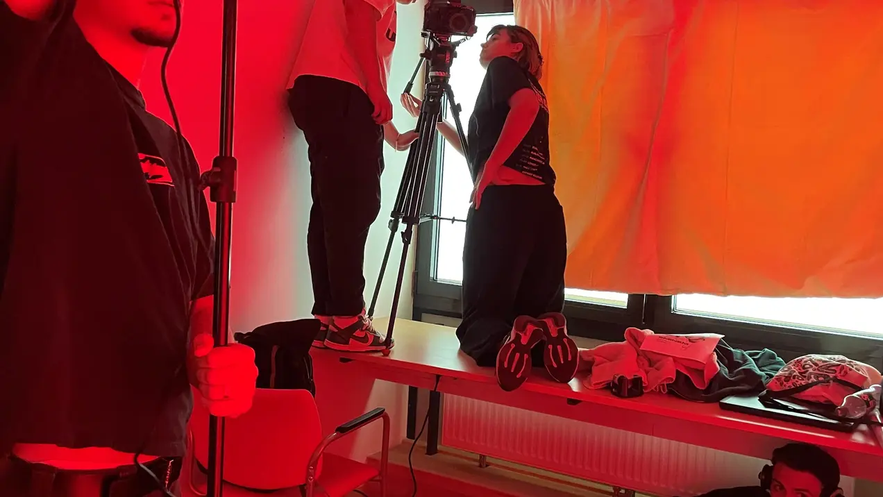 Eine Gruppe von vier Personen arbeitet in einem Raum mit rotem Licht. Zwei Personen befinden sich auf einem Tisch, eine filmt mit einer Kamera, während die andere hilft. Eine Person hält ein Licht, und eine andere liegt auf dem Boden und bereitet etwas vor.