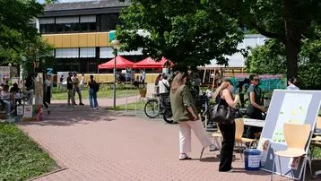 Auf dem Bild ist ein lebhafter Außenbereich einer Veranstaltung zu sehen, mit mehreren Ständen, Menschen, die sich unterhalten und eine grüne Umgebung. Es gibt Zelte, Tische und Sitzgelegenheiten, die auf eine gemeinschaftliche Aktivität hindeuten. Die Szene vermittelt eine gesellige, einladende Atmosphäre.
