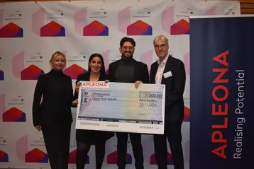 Die vier Personen auf dem Bild stehen zusammen und halten einen großen Scheck über 1.000 Euro. In der Mitte stehen Ilyas Baladi, der Gewinner des Apleona-Förderpreises. Links daneben befindet sich Alumna Daniela Ay, die den Scheck übergibt.