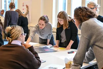 A group of female students are working together at a table. They are discussing and looking at plans. One woman is pointing at a document while others listen attentively and take notes. The room has large windows that let in natural light.