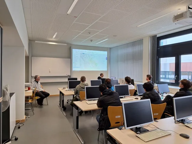 In einem Seminarraum sitzen Schülerinnen und Schüler an Computern, während ein Lehrer vorne eine Präsentation mit einer Karte an der Wand zeigt. Die Schüler:innen sind aufmerksam und folgen den Erklärungen. Das Licht ist hell und die Fenster lassen Tageslicht herein.