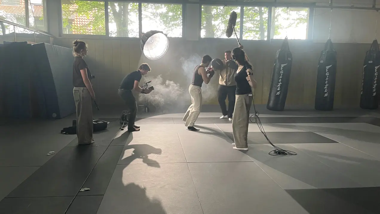 Eine Gruppe von fünf Personen filmt in einem Trainingsraum, der mit Boxsäcken ausgestattet ist. Zwei Personen trainieren gemeinsam, während die anderen mit Kamera und Lichttechnik arbeiten. Der Raum wirkt dynamisch und ist teilweise durch eine Nebelmaschine eingehüllt.