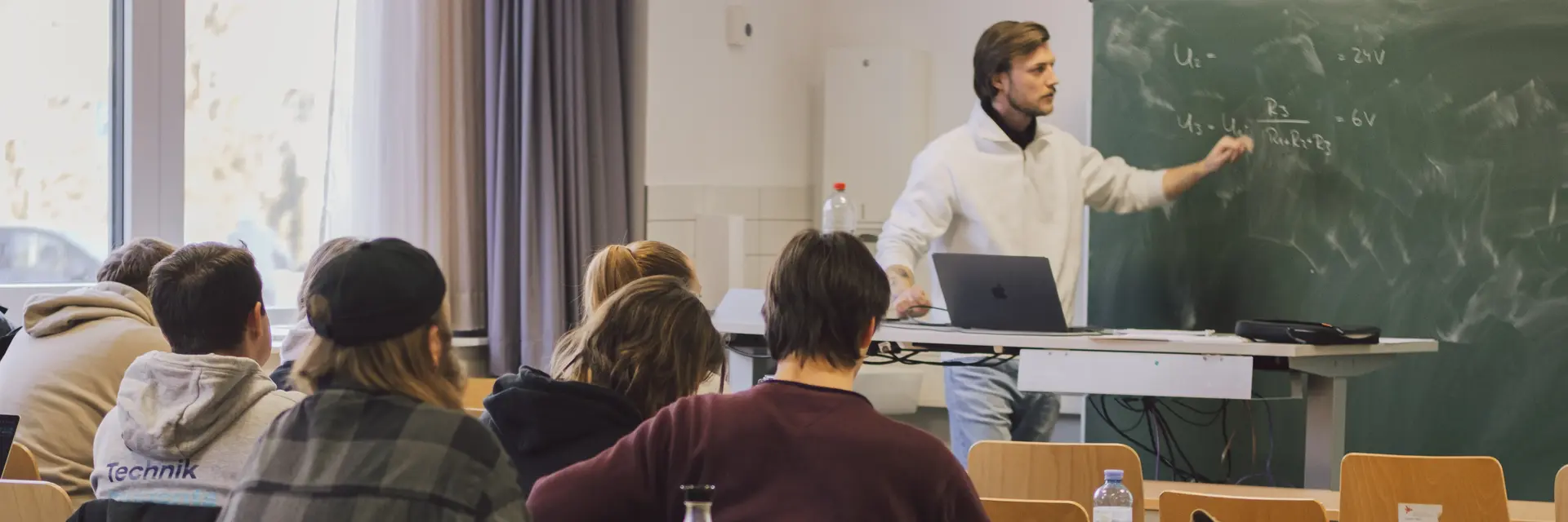 Ein Dozent steht vor einer Tafel und erklärt den Schülern in einem Klassenzimmer. Die Studierenden sitzen an Tischen, einige nutzen Laptops. Helle Fenster sorgen für Tageslicht im Raum.