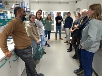 Eine Gruppe von zehn Personen steht in einem Labor. Ein Mann erklärt etwas, während die anderen aufmerksam zuhören. Die Atmosphäre ist informell und kooperativ, mit Laborausrüstung im Hintergrund.