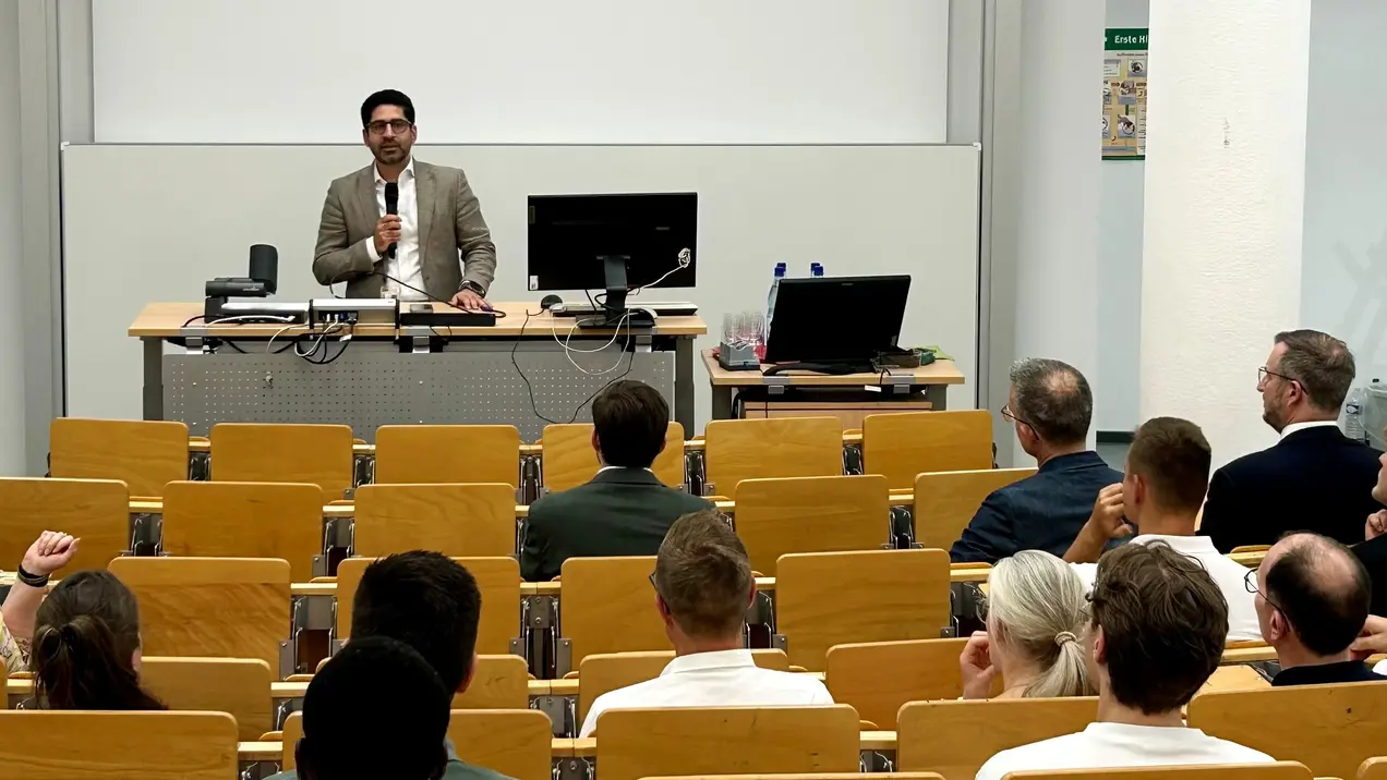 Ein Mann in Anzug steht am Pult und spricht zu einem Publikum von etwa zehn Personen, die in einem Seminarraum sitzen. Der Raum ist mit Holztischen und Stühlen ausgestattet. Im Hintergrund sind ein Computer und Präsentationsmaterialien sichtbar.