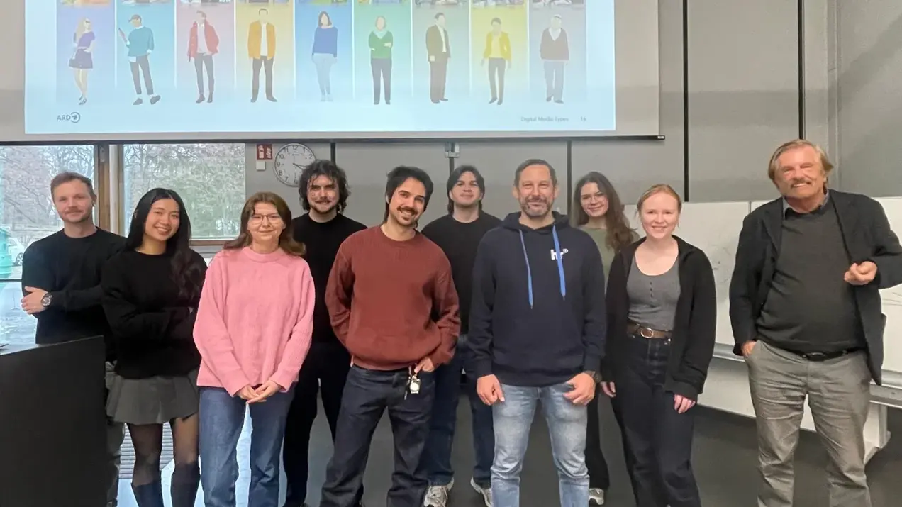 Eine Gruppe von zehn Personen steht nebeneinander in einem modernen Raum. Im Hintergrund ist eine Präsentation zu sehen, die verschiedene digitale Medientypen zeigt. Die Personen lächeln und wirken freundlich, während sie in die Kamera schauen.