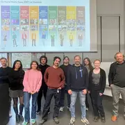 Eine Gruppe von zehn Personen steht nebeneinander in einem modernen Raum. Im Hintergrund ist eine Präsentation zu sehen, die verschiedene digitale Medientypen zeigt. Die Personen lächeln und wirken freundlich, während sie in die Kamera schauen.