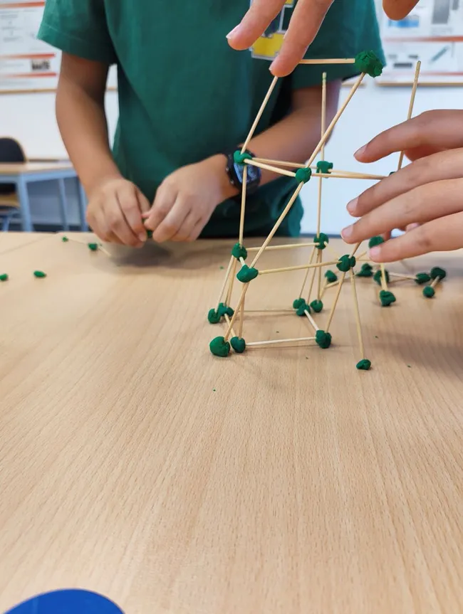 Zwei Hände arbeiten an einem Modell aus Zahnstocher und Knete. Eine Hand hält einen Zahnstocher, während mit der anderen ein grüner Kneteball auf einen weiteren Zahnstocher gesetzt wird. Im Hintergrund sind verschwommene Schulmöbel zu sehen.