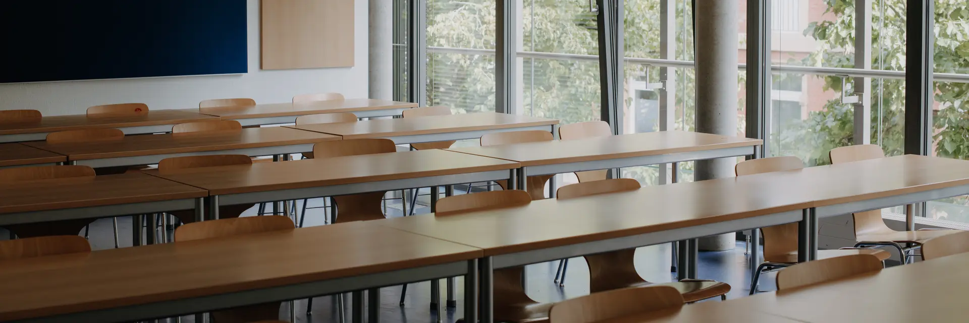 Ein heller Klassenraum mit mehreren langen Tischen und Stühlen. An einer Wand befindet sich eine schwarze Tafel. Große Fenster bieten Blick auf grüne Bäume im Freien. Der Raum wirkt freundlich und einladend.