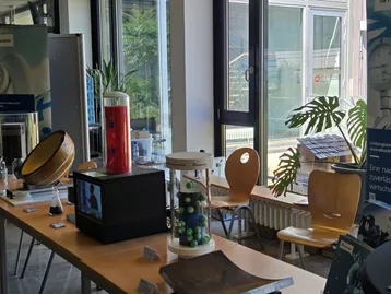 Der Bildinhalt zeigt einen Ausstellungsbereich mit verschiedenen Objekten. Auf einem Tisch sind ein schwarzer Monitor, ein Behälter mit grünen Kugeln und ein rotes Zylindermodell zu sehen. Im Hintergrund sind Pflanzen und Stühle sichtbar, das Licht fällt durch große Fenster.