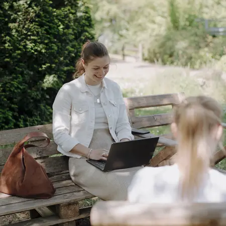 Eine lächelnde Frau sitzt auf einer Bank in einem Garten und arbeitet am Laptop. Sie trägt eine weiße Jacke und hat eine braune Tasche neben sich. Im Hintergrund sind grüne Pflanzen und Wiesen zu sehen. Eine zweite Person sitzt mit dem Rücken zur Kamera.