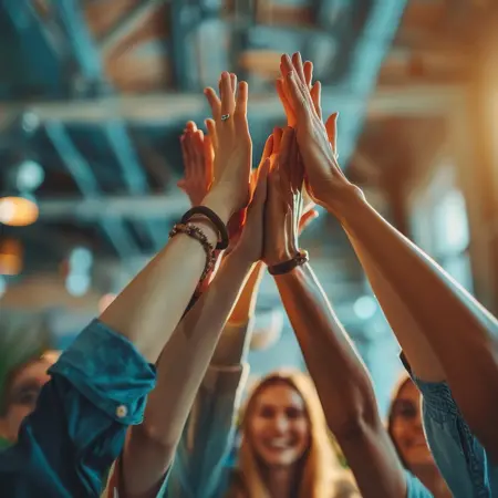 Eine Gruppe von Menschen hebt freudig ihre Hände in die Höhe, um sich gegenseitig zu gratulieren oder einen Erfolg zu feiern. Im Hintergrund sind unscharfe Gesichter und eine moderne, helle Umgebung mit Pflanzen und großen Fenstern zu sehen.