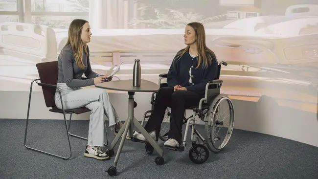 Zwei Personen sitzen an einem Tisch vor einer Leinwand mit einem Krankenhaus als Hintergrund. Die Person rechts sitzt in einem Rollstuhl und die Person links schreibt sich Sachen auf einem Block auf.