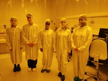 Fünf Personen in Schutzkleidung, einschließlich Hauben und Mundschutz, stehen nebeneinander in einem Labor mit gelblichem Licht. Alle tragen weiße Kittel und spezielle Schuhe, was auf eine sterile Umgebung hinweist.