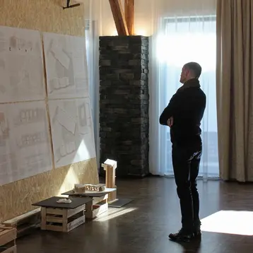 Ein Mann steht vor einer Wand, die mit Plänen und Zeichnungen bedeckt ist. Er betrachtet die Arbeiten aufmerksam, in einem Raum mit Holzelementen und großen Fenstern, die Tageslicht einlassen. Unter den Postern befinden sich Holzkisten auf denen Architekturmodelle stehen.