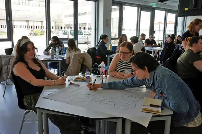 Eine Gruppe von Menschen sitzt an Tischen in einem modernen Raum. Einige Teilnehmer arbeiten gemeinsam an großen Papierbögen, zeichnen Diagramme und schreiben Notizen. Die Atmosphäre ist kreativ und kooperativ, mit Getränken auf den Tischen.