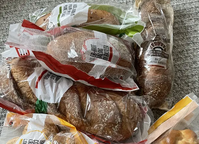 Eine Ansammlung von verschiedenen Brotsorten, verpackt in Plastiktüten. Dazu gehören Brötchen, eine Art Zopf und Kekse. Die Tüten sind teils durchsichtige Folien, die den Inhalt sichtbar machen. Die Brote liegen zufällig auf einem hellen Untergrund.