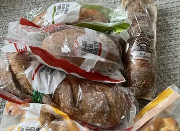 Eine Ansammlung von verschiedenen Brotsorten, verpackt in Plastiktüten. Dazu gehören Brötchen, eine Art Zopf und Kekse. Die Tüten sind teils durchsichtige Folien, die den Inhalt sichtbar machen. Die Brote liegen zufällig auf einem hellen Untergrund.