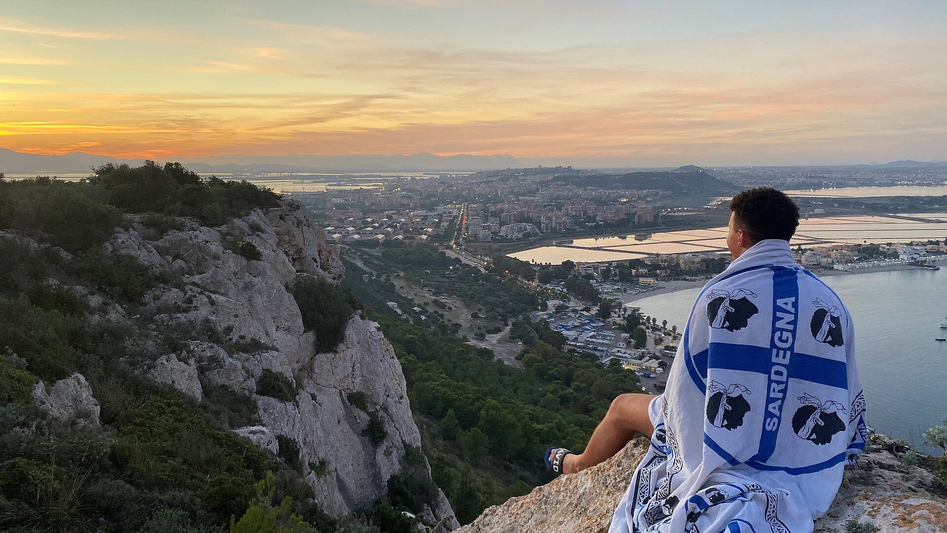 Ouassim Lamrini Ouassim Lamrini sitzt auf einem Felsen, er trägt eine Flagge Sardiniens über der Schulter und blickt in den Sonnenuntergang