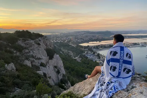 Ouassim Lamrini sitzt auf einem Felsen, er trägt eine Flagge Sardiniens über der Schulter und blickt in den Sonnenuntergang