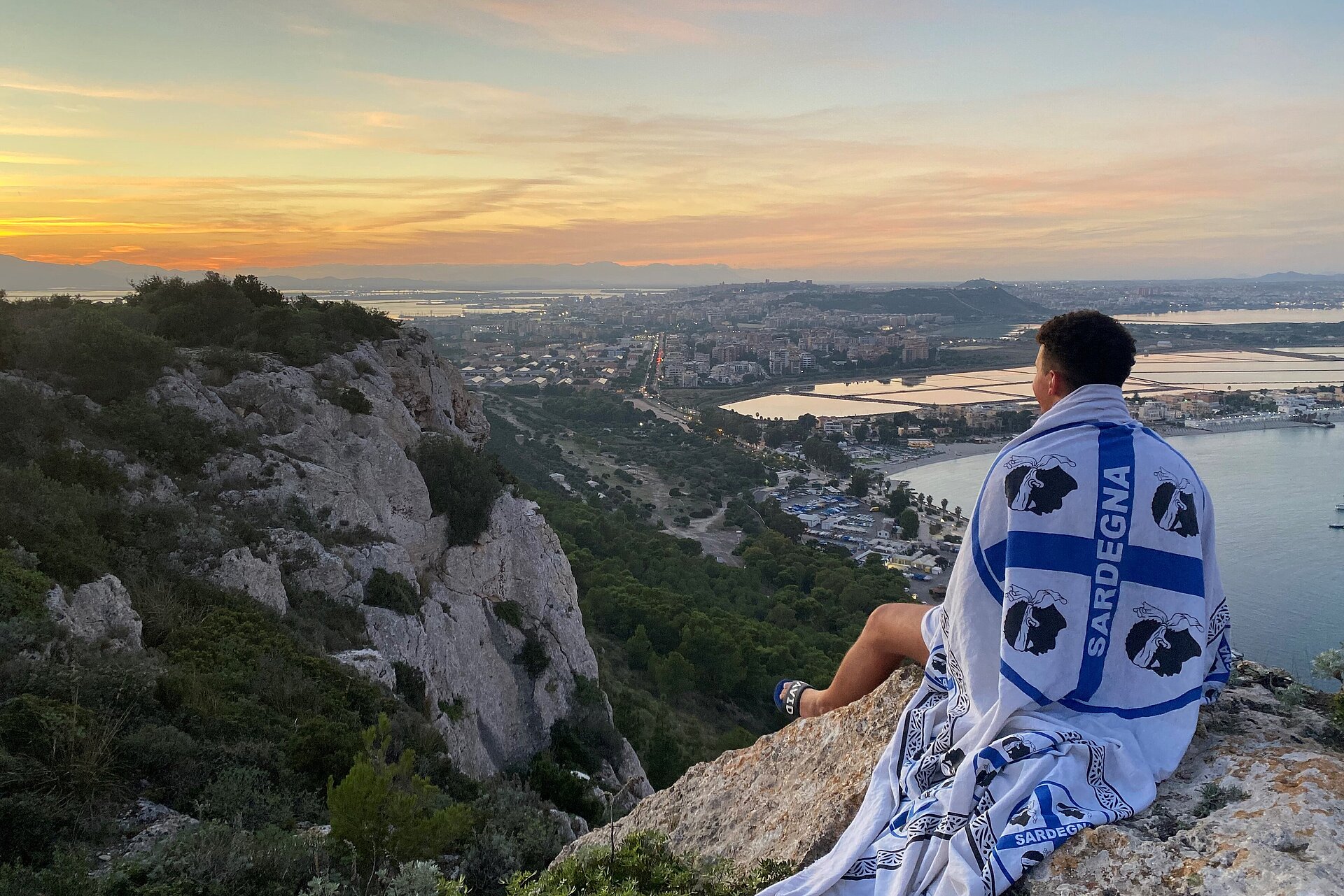Ouassim Lamrini Ouassim Lamrini sitzt auf einem Felsen, er trägt eine Flagge Sardiniens über der Schulter und blickt in den Sonnenuntergang