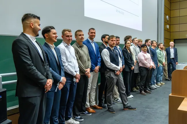Eine Gruppe von Männern und einer Frau steht auf einer Bühne in einem Veranstaltungsraum, gekleidet in Anzügen und formeller Kleidung. Sie präsentieren sich lächelnd vor einem Publikum. Im Hintergrund ist ein Bildschirm zu sehen. Das Bild vermittelt einen festlichen Anlass, wahrscheinlich eine Abschlussfeier oder Ehrungsveranstaltung.