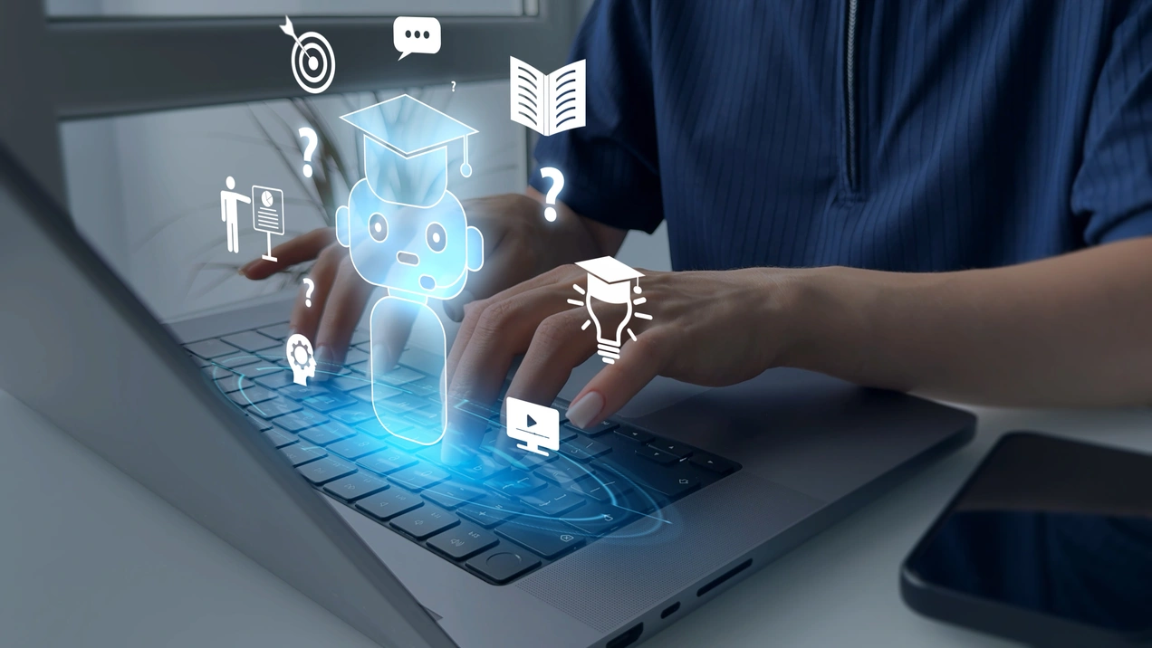Eine Person tippt auf einer Laptop-Tastatur. Über der Tastatur sind digitale Symbole von einem Roboter, einem Buch, einer Glühbirne und Fragezeichen zu sehen, die Lernprozesse und digitale Interaktion darstellen.