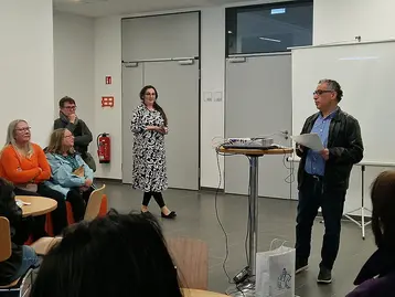 Eine Gruppe von Menschen sitzt an Tischen und hört einem Mann zu, der am Pult spricht. Im Hintergrund stehen eine Frau in einem gemusterten Kleid und eine weitere Person. Die Szene findet in einem hellen Raum statt, der für eine Präsentation vorbereitet ist.