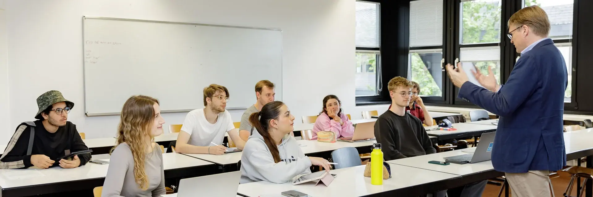 Ein Professor steht an der Vorderseite eines Raums der Hochschule RheinMain und spricht mit einer Gruppe von acht Studierenden, die an Tischen sitzen. Einige der Studierenden nutzen Laptops. Die Atmosphäre wirkt engagiert und interaktiv.