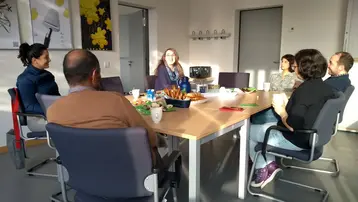 Ein Treffen in einem Konferenzraum mit sechs Personen, die um einen Tisch sitzen. In der Mitte des Tisches befinden sich Snacks und Getränke. Eine Person spricht, während die anderen aufmerksam zuhören. Tageslicht fällt durch ein Fenster in den Raum.