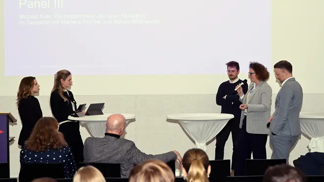 Michael Auer, Pia Hoppenberg, Leon Kesselhof, Marianne Romba und Natalie Wisniewski stehen an Stehtischen und diskutieren. Im Hintergrund sind Zuhörer zu sehen, die aufmerksam zuschauen. Ein Bildschirm zeigt den Titel „Panel III“ des 2. Wiesbadener Tag für sozial nachhaltiges Investieren an der Hochschule RheinMain.