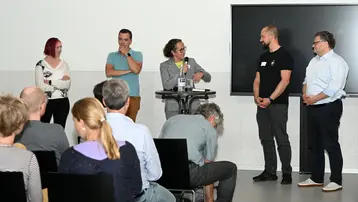 Eine Gruppe von fünf Personen steht auf einer Bühne während einer Veranstaltung. Eine Person spricht ins Mikrofon, während die anderen aufmerksam zuhören. Im Hintergrund sind Stühle mit Publikum zu sehen.