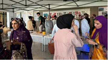 Die Szene zeigt eine belebte Veranstaltung unter einem Zelt. Mehrere Menschen, hauptsächlich Frauen in traditionellen Kleidern, unterhalten sich, während im Hintergrund eine Gruppe von Teilnehmern steht. Tische mit Speisen und Informationsmaterial sind ebenfalls zu sehen.