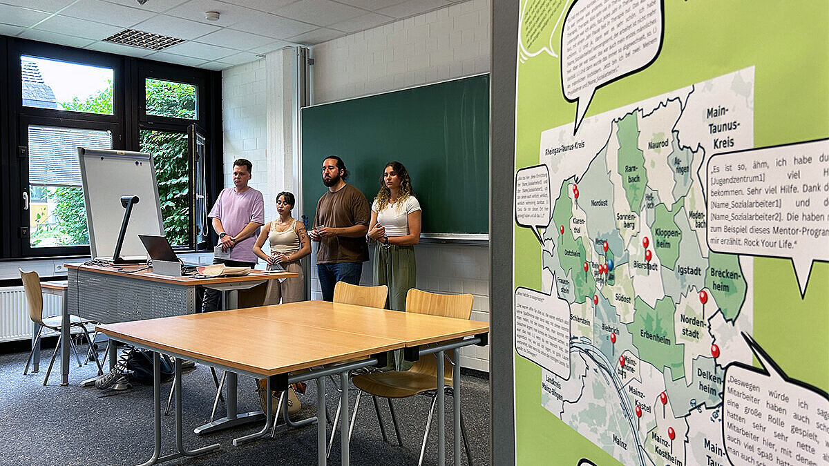 Vier junge Menschen stehen während einer Präsenation vor einer Tafel in einem Seminarraum, rechts daneben ist eine Stellwand mit einer Karte und Sprechblasen zu sehen.