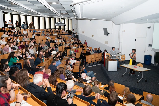 Ein gut besuchter Hörsaal mit zahlreichen Zuhörern. Auf der Bühne präsentieren zwei Personen, während das Publikum aufmerksam zuhört und applaudiert. Die Atmosphäre ist interaktiv und gespannt, was auf ein wichtiges Thema oder eine Präsentation hinweist.