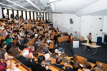 Ein gut besuchter Hörsaal mit zahlreichen Zuhörern. Auf der Bühne präsentieren zwei Personen, während das Publikum aufmerksam zuhört und applaudiert. Die Atmosphäre ist interaktiv und gespannt, was auf ein wichtiges Thema oder eine Präsentation hinweist.