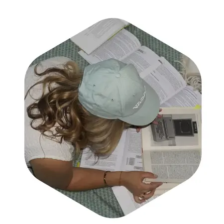 Eine Person mit lockigem Haar und einer blauen Kappe liegt auf dem Boden und studiert mehrere aufgeschlagene Bücher. Sie hält ein Buch in der Hand und scheint konzentriert zu lesen. Die Umgebung ist mit einem Teppich ausgelegt.