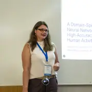 Eine Präsentation wird von einer Frau gehalten, die einen weißen Oberteil und eine blaue Halskette trägt. Sie steht vor einer Leinwand mit einem Text über ein neuronales Netzwerk zur Erkennung menschlicher Aktivitäten. Die Umgebung ist hell und professionell.
